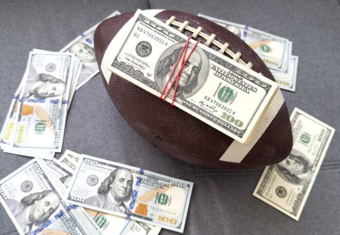 American,Football,Or,Rugby.,Dollars.,Concept,Sport,And,Money.,Sports Football with a stack of U.S. hundred-dollar bills secured by rubber bands