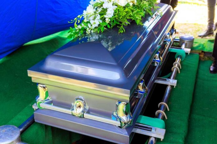 Funeral,Silver,Casket,As,Decorated,Coffin,Is,Lowered,At,During A silver casket adorned with flowers at a gravesite
