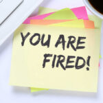 A sticky note with the message 'YOU ARE FIRED!' placed on a desk next to a keyboard and a cup of coffee