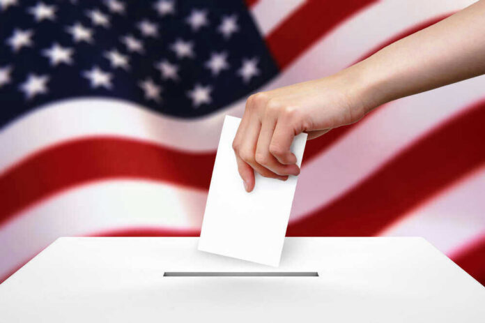 shutterstock_99507023 Hand dropping ballot into box, American flag background.