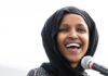 Omar’s Home Raided Ilhan Omar with a black headscarf smiles near a microphone.