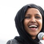 Omar’s Home Raided Ilhan Omar with a black headscarf smiles near a microphone.