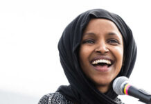 Omar’s Home Raided Ilhan Omar with a black headscarf smiles near a microphone.