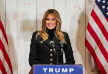 Melania Drops WAR Bombshell Woman speaking at Trump podium, two American flags present.